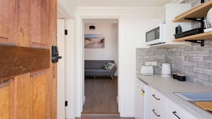 A modern kitchen with white cabinets, a grey countertop, and a microwave. There is a grey sofa in the adjacent living area with a beach-themed picture on the wall.