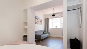 A bedroom with a bed, a grey sofa, a window with curtains, a hanging light fixture, and a small black bag on a shelf.
