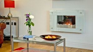 Uma sala de estar moderna com uma mesa de centro de vidro, um vaso com flores, um abajur vermelho, uma TV de tela plana e uma lareira.