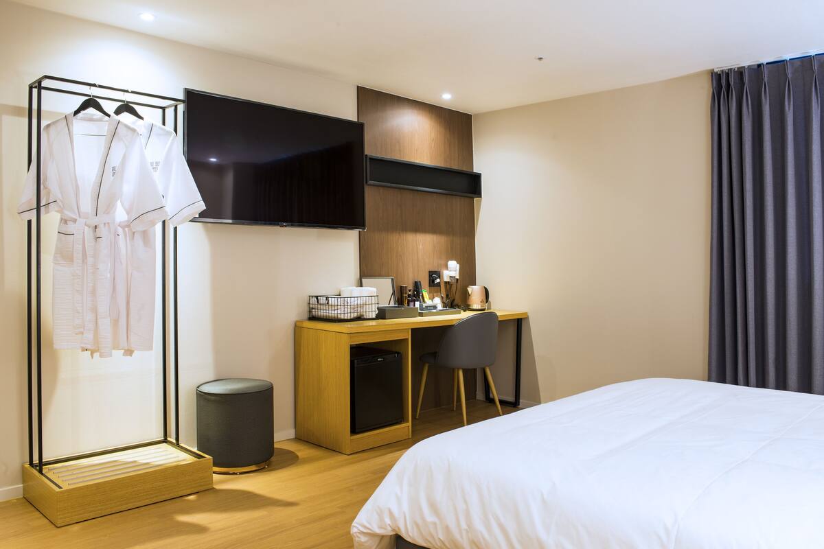 A hotel room with a bed, a desk with a chair, a TV mounted on the wall, and a clothing rack with robes.