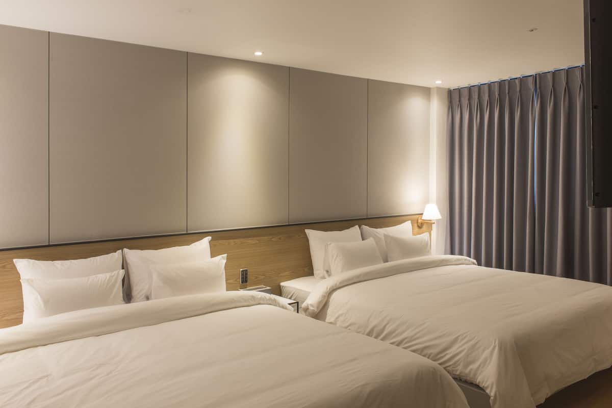 A hotel room with two beds, a wooden headboard, a bedside table with a lamp, and a view of the city through curtains.