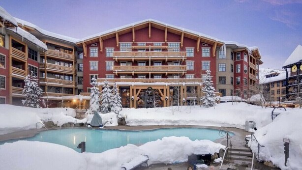 A hotel with a pool surrounded by snow, featuring multiple balconies and a large building facade. - The Village At Mammoth, 2BR/2B Suite, Great Location Above Shops & Restaurants   (Mammoth Lakes)