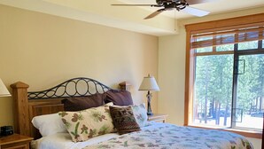 A bedroom with a bed, wooden floor, ceiling fan, bedside table with lamp, and a window with a view of trees. - The Village At Mammoth, 2BR/2B Suite, Great Location Above Shops & Restaurants   (Mammoth Lakes)