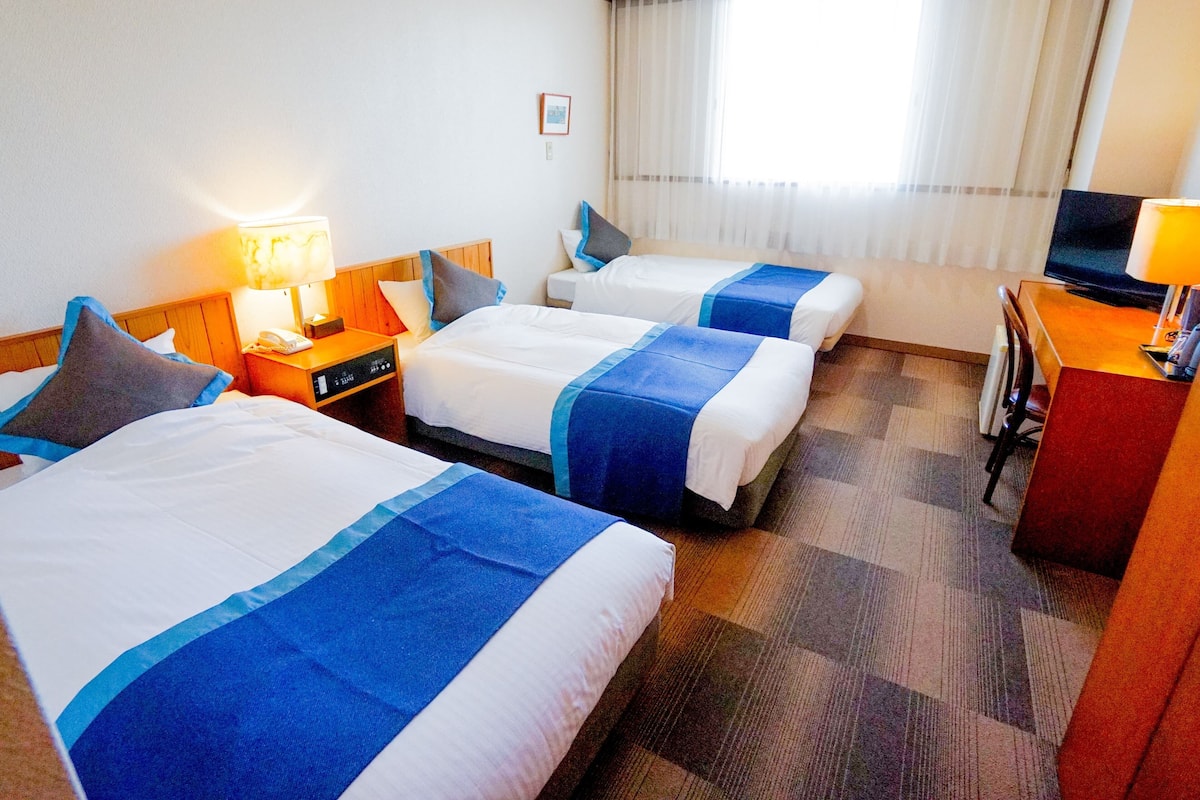 Quarto de hotel com três camas, uma escrivaninha, televisão e janela com cortinas.