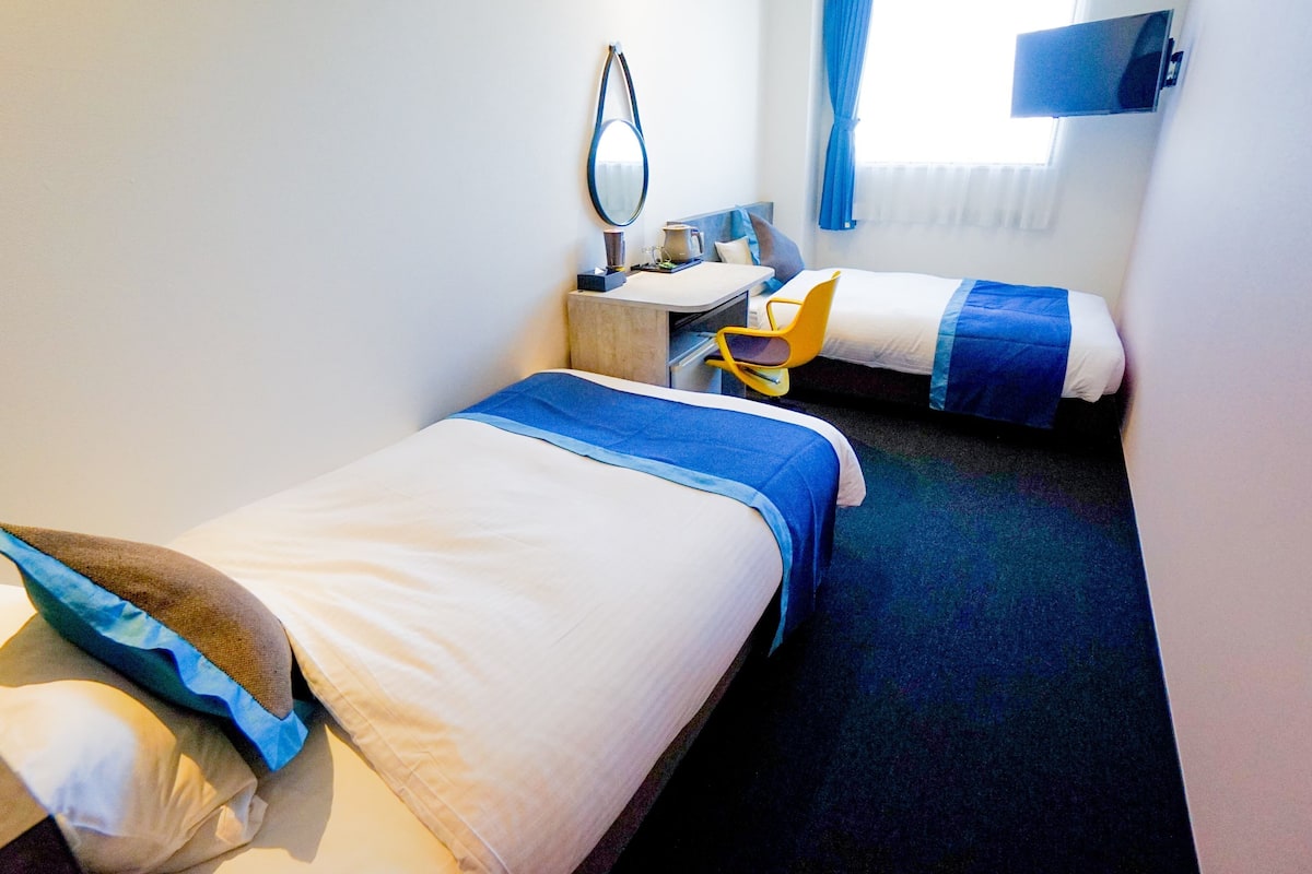 Quarto de hotel com duas camas, uma mesa, uma cadeira, uma televisão e uma janela com cortinas.