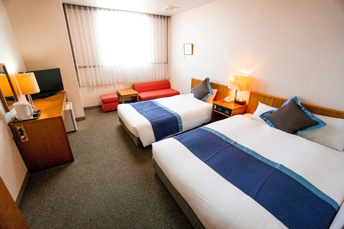 Quarto de hotel com duas camas, uma televisão, uma escrivaninha com candeeiro e um sofá vermelho, além de uma janela com cortinas.