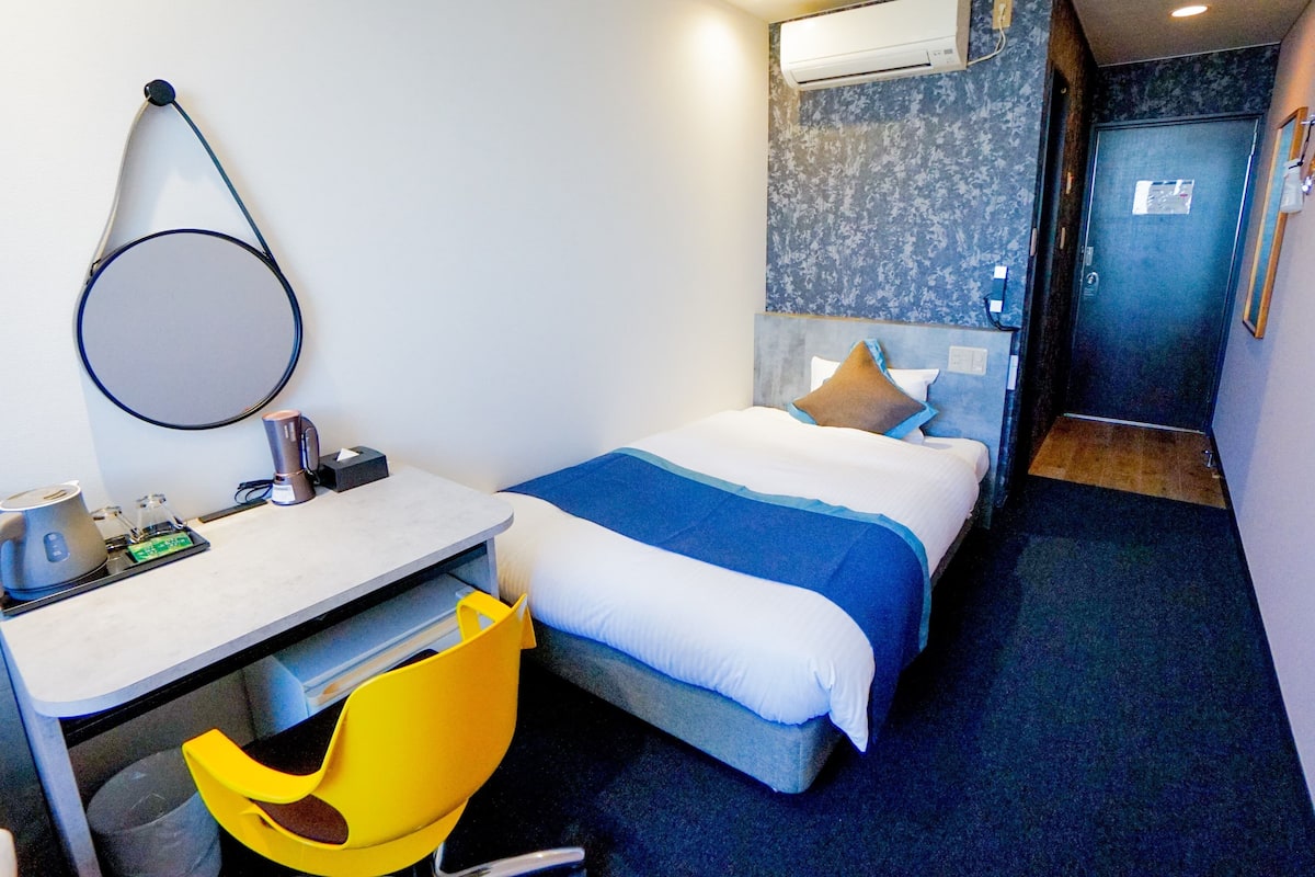 Quarto de hotel pequeno com uma cama, uma escrivaninha e uma cadeira.