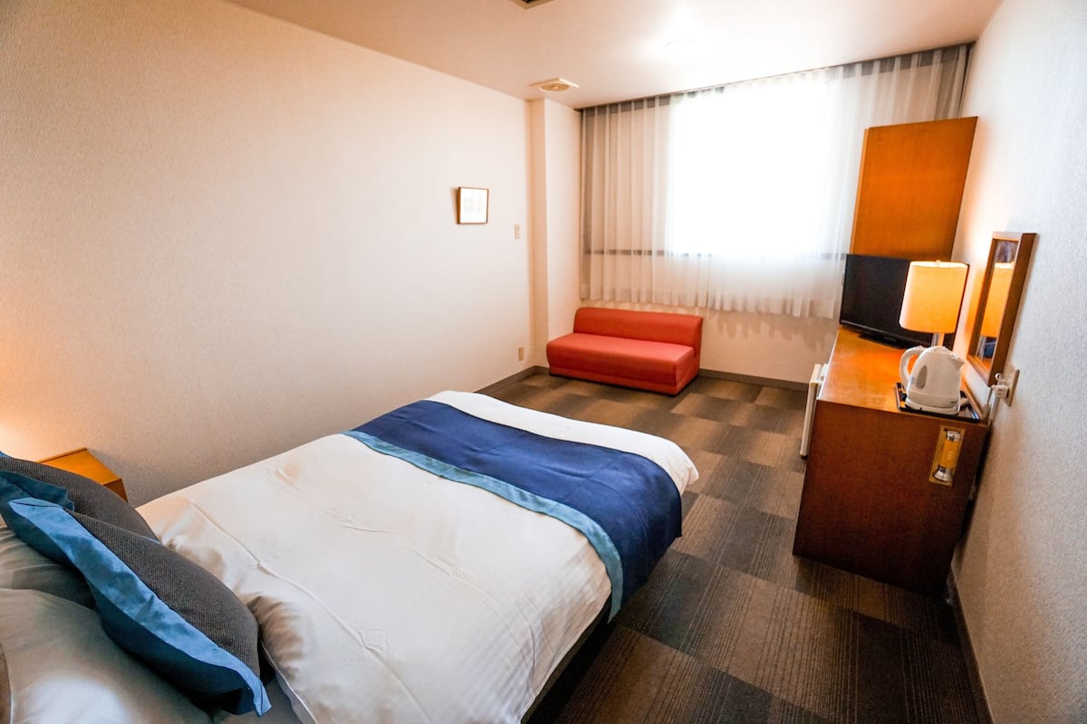 Quarto de hotel com cama, uma cadeira vermelha, escrivaninha com abajur e televisão.