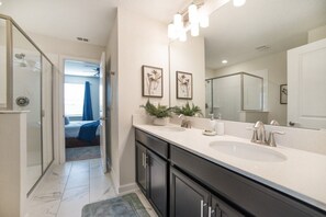 Une salle de bain moderne avec un grand meuble-lavabo, deux vasques et une douche. Des tableaux encadrés ornent le mur, et une plante en pot est posée sur le meuble-lavabo.