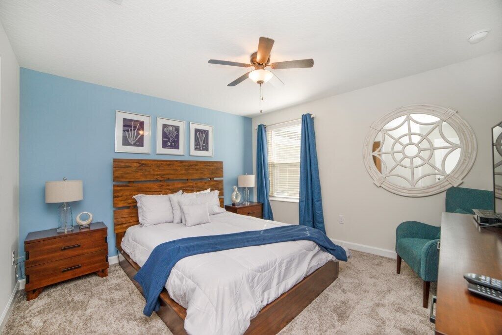 A bedroom with a wooden headboard, a bed with white bedding, a ceiling fan, a round decorative window, and a nightstand with a lamp.