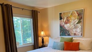 A bedroom with a large bed, a nightstand with a lamp, a window with curtains, and a floral painting on the wall. - Gracie's Homer House; Home With A View (Homer)