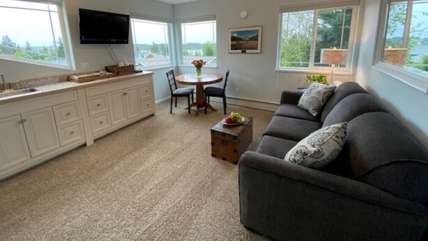 A modern kitchen with white cabinets, a dining area with a round table and chairs, a gray sofa with patterned cushions, and large windows offering views of greenery. - Gracie's Homer House; Home With A View (Homer)