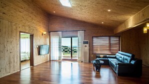 A spacious room with wooden walls, a black leather sofa, and a large sliding glass door leading to a balcony. - Witch's  broomstick Pension (Jeju City)