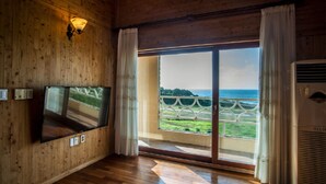 A room with wooden walls, a flat-screen TV mounted on the wall, a large sliding glass door leading to a balcony with a view of the sea and greenery. - Witch's  broomstick Pension (Jeju City)