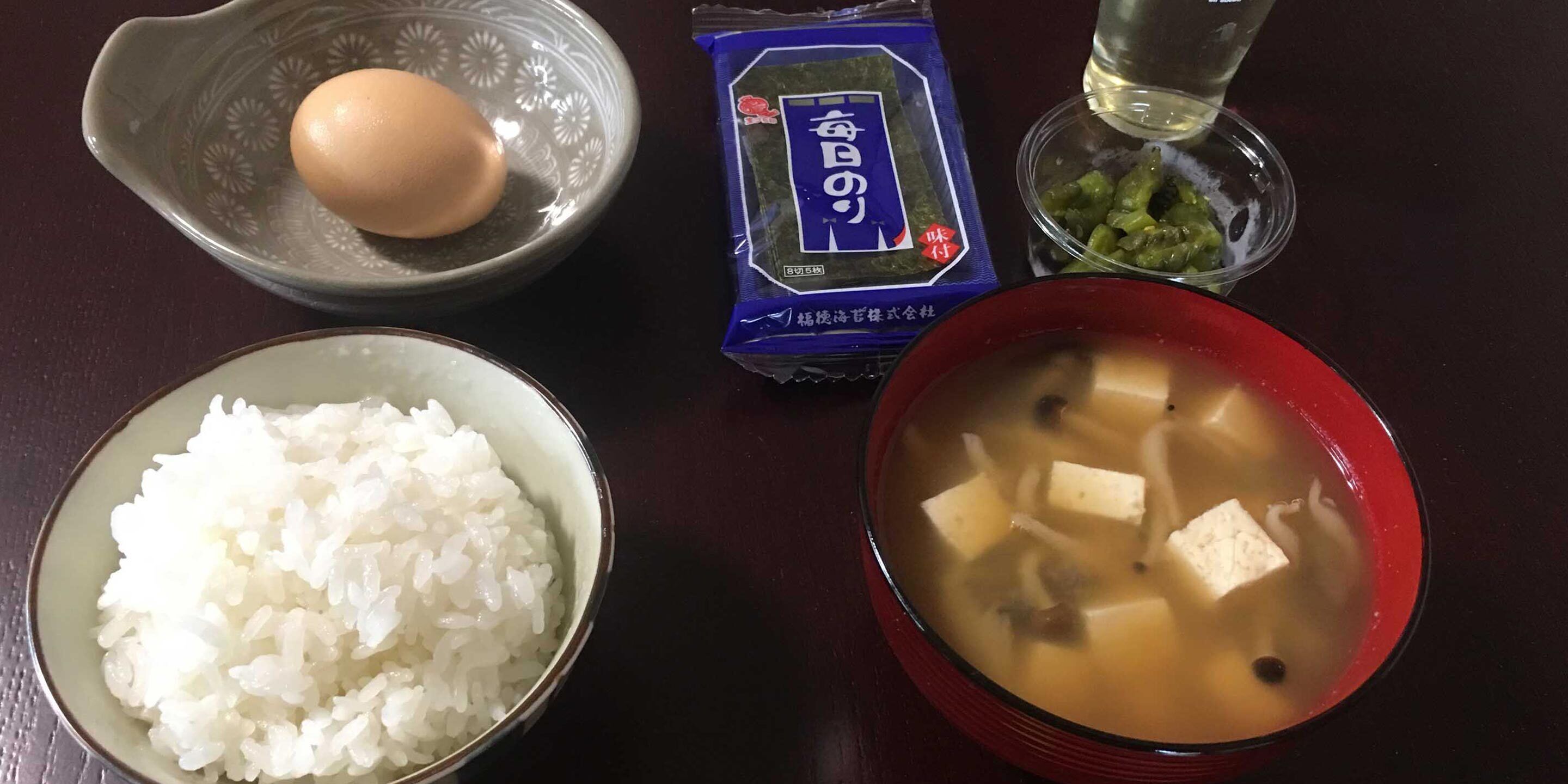 Una mesa con un tazón de arroz blanco, un tazón de sopa de tofu, un paquete de algas marinas y un vaso de agua, junto con un huevo crudo.