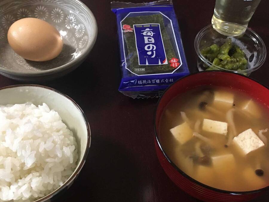 Una mesa con un tazón de arroz blanco, un tazón de sopa de tofu, un paquete de algas marinas y un vaso de agua, junto con un huevo crudo.