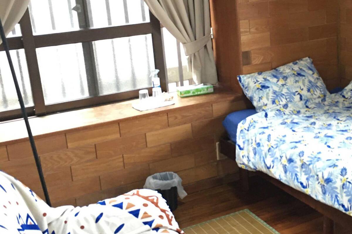 Een kleine kamer met een enkel bed, een houten hoofdbord, een raam met gordijnen, een nachtkastje met toiletartikelen en een prullenbak.