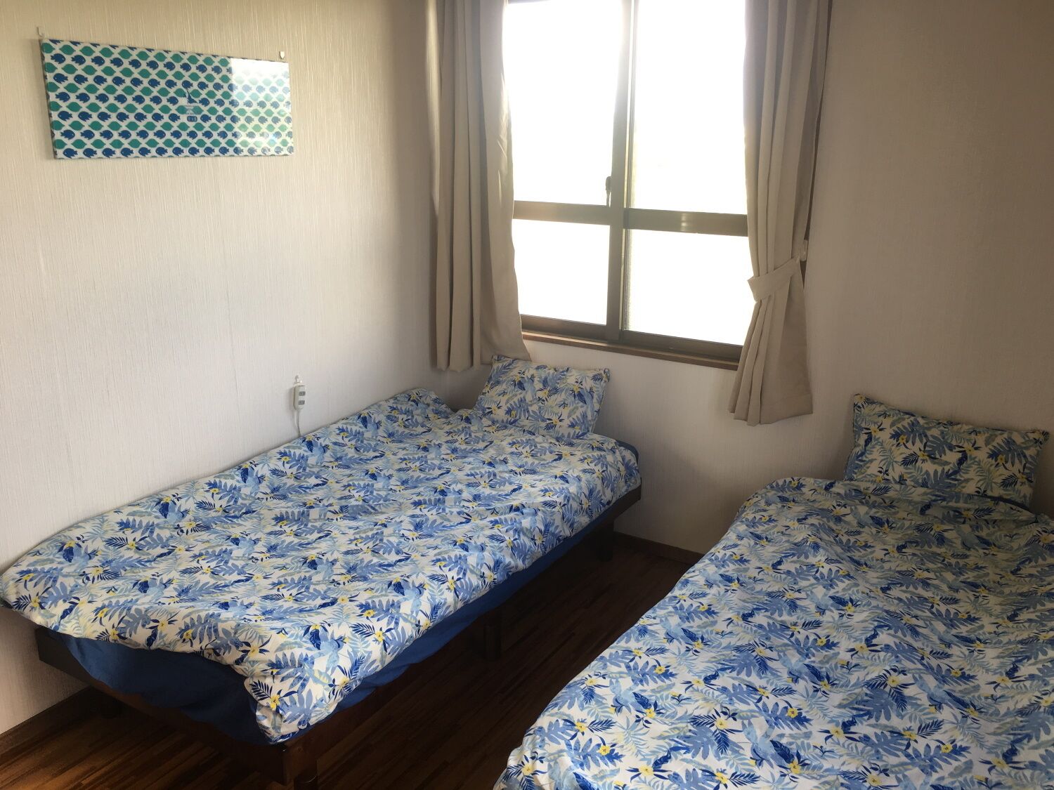 Una habitación con dos camas individuales, una ventana con cortinas, un interruptor de luz en la pared y un tapiz decorativo en la pared.