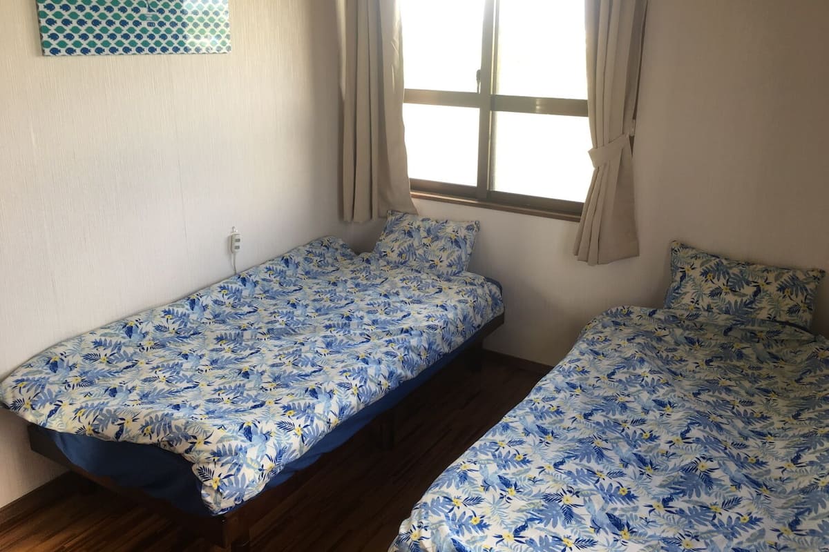 Een kamer met twee éénpersoonsbedden, een raam met gordijnen, een lichtschakelaar aan de muur en een wanddecoratie met een patroon.