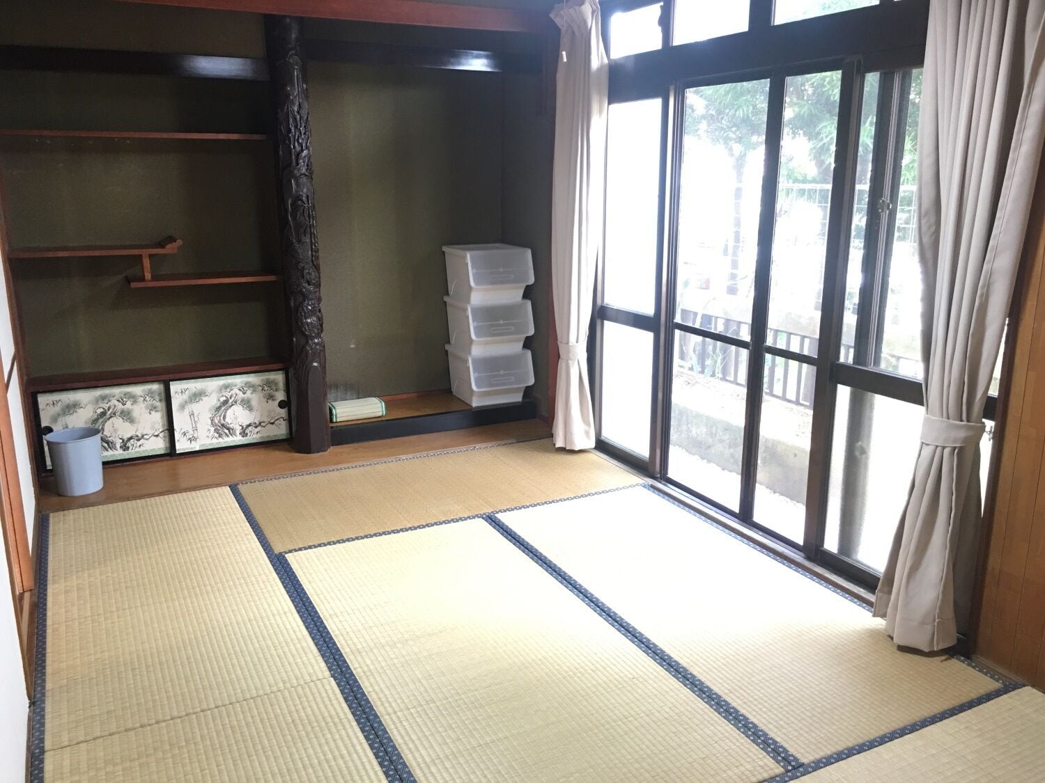 A room with tatami flooring, a sliding door, a window with curtains, a storage unit, and a small trash bin.
