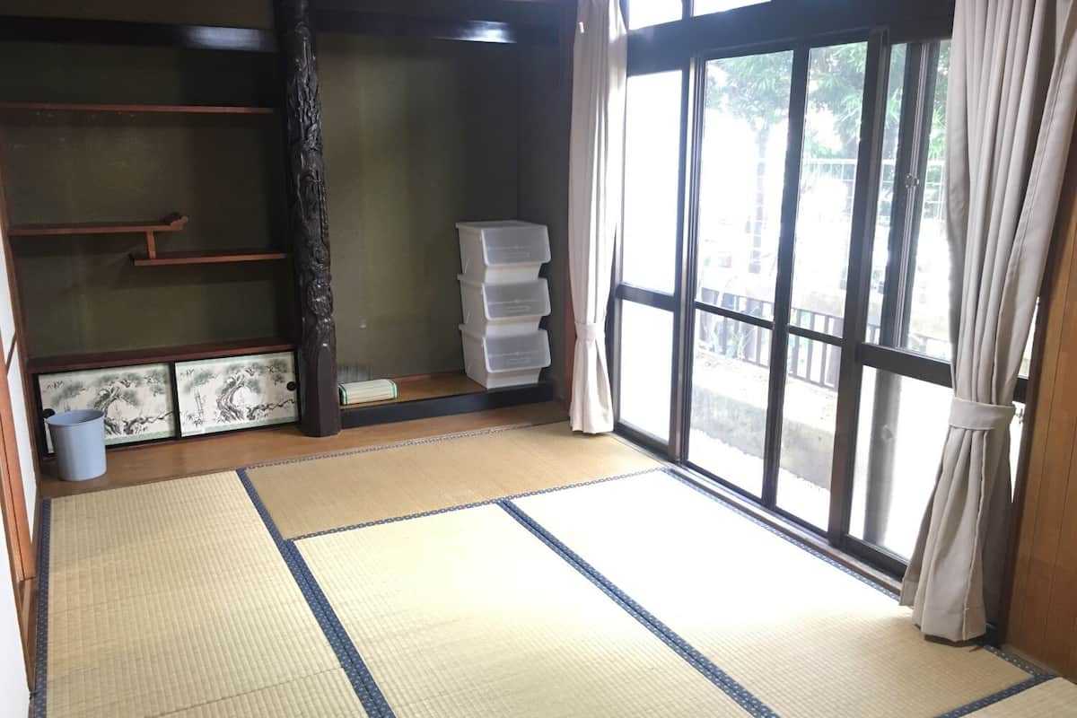 Een kamer met tatami vloerbedekking, een schuifdeur, een raam met gordijnen, een opbergruimte en een kleine prullenbak.