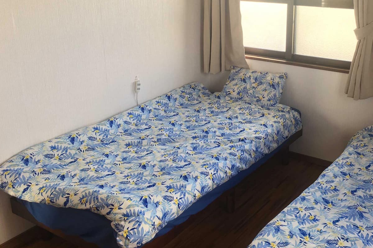 Een kamer met twee eenpersoonsbedden, elk met een bloemenprint sprei. Er is een raam met gordijnen, en een stopcontact is zichtbaar.