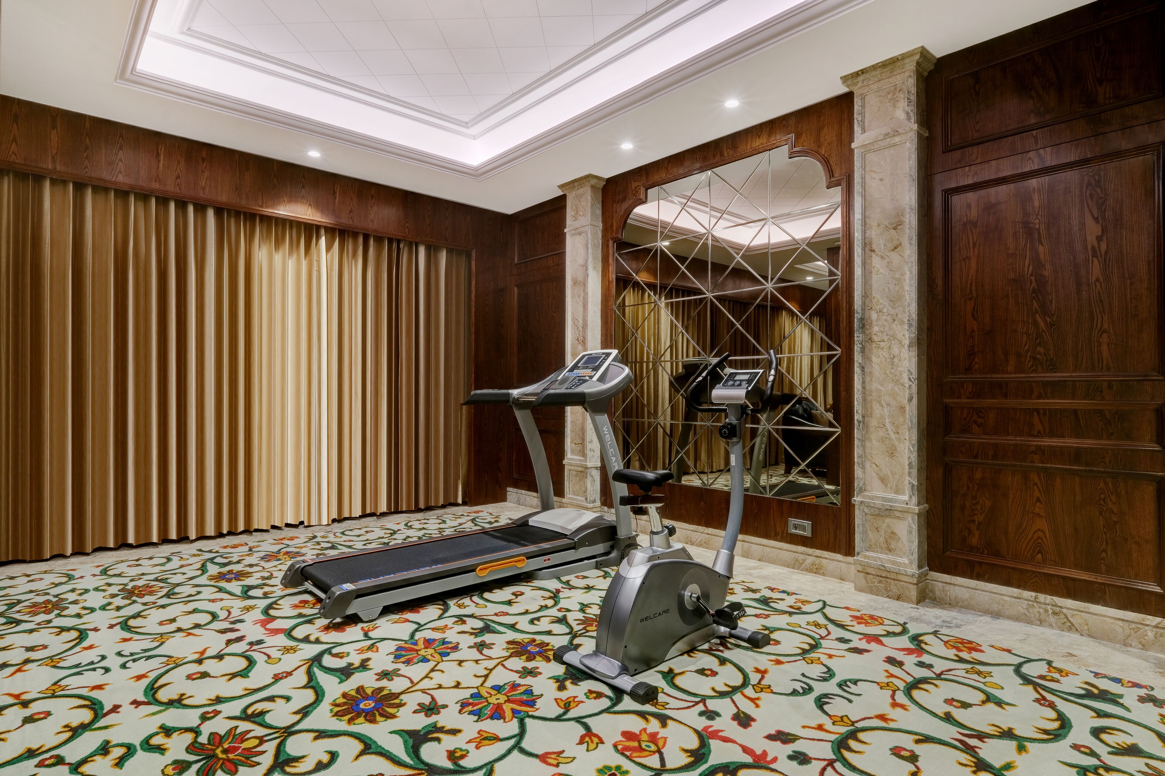 Una habitación con cinta de correr y bicicleta estática, alfombra estampada, paneles de madera y un espejo grande.