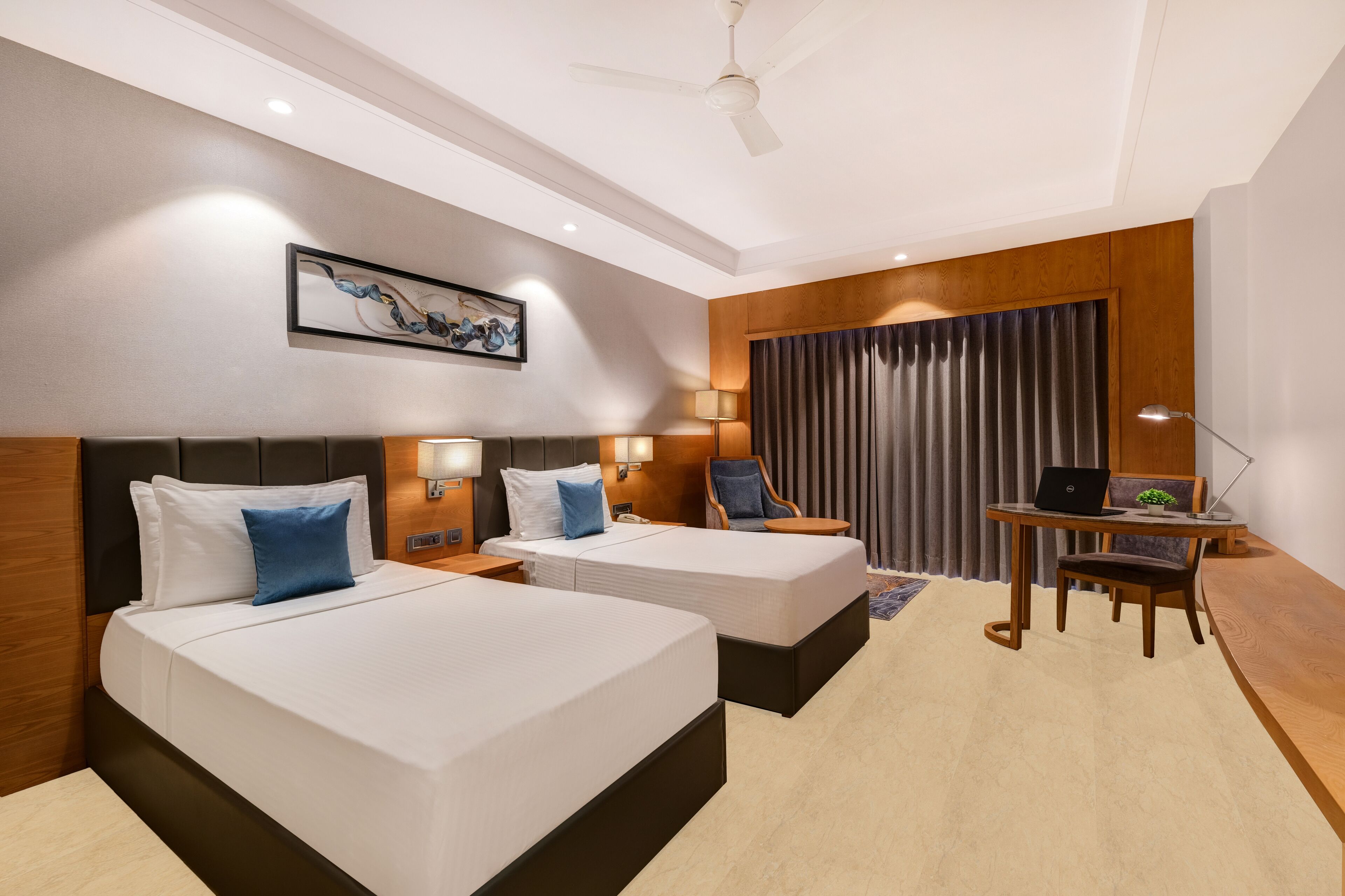 Une chambre d’hôtel avec deux lits, un bureau avec un ordinateur portable, une chaise et un ventilateur de plafond.