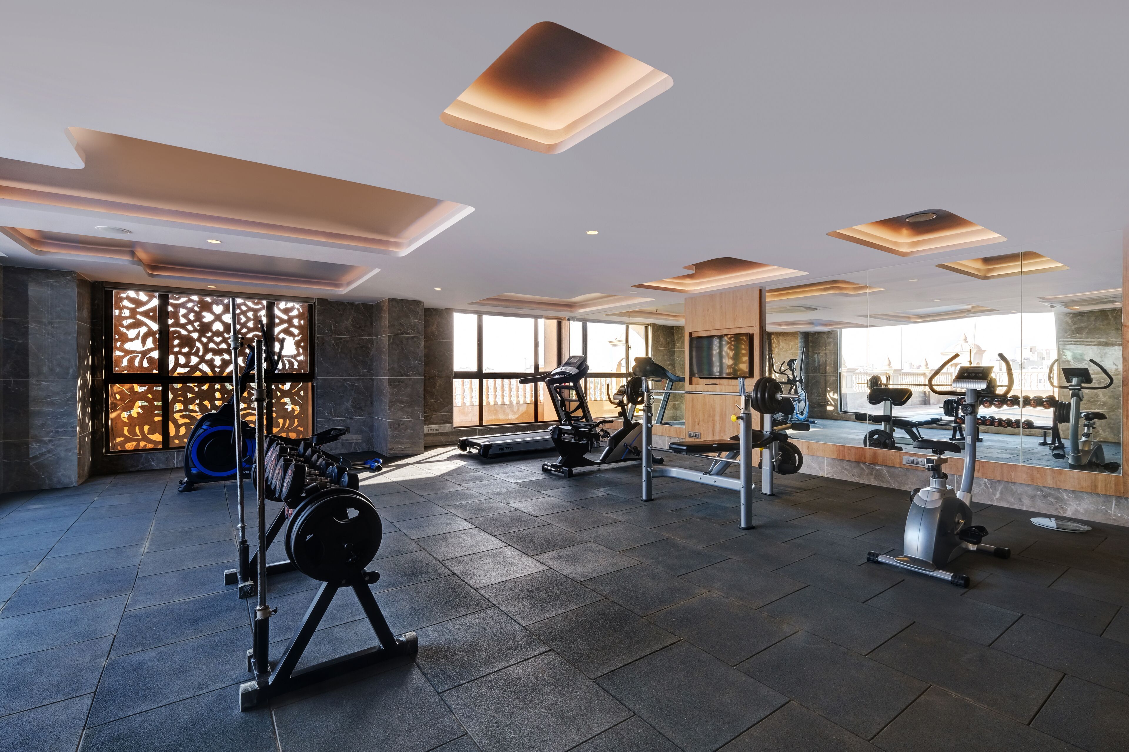 Une salle de sport moderne dotée de divers appareils d’exercice, de grandes fenêtres et d’un plafond haut avec des luminaires encastrés.