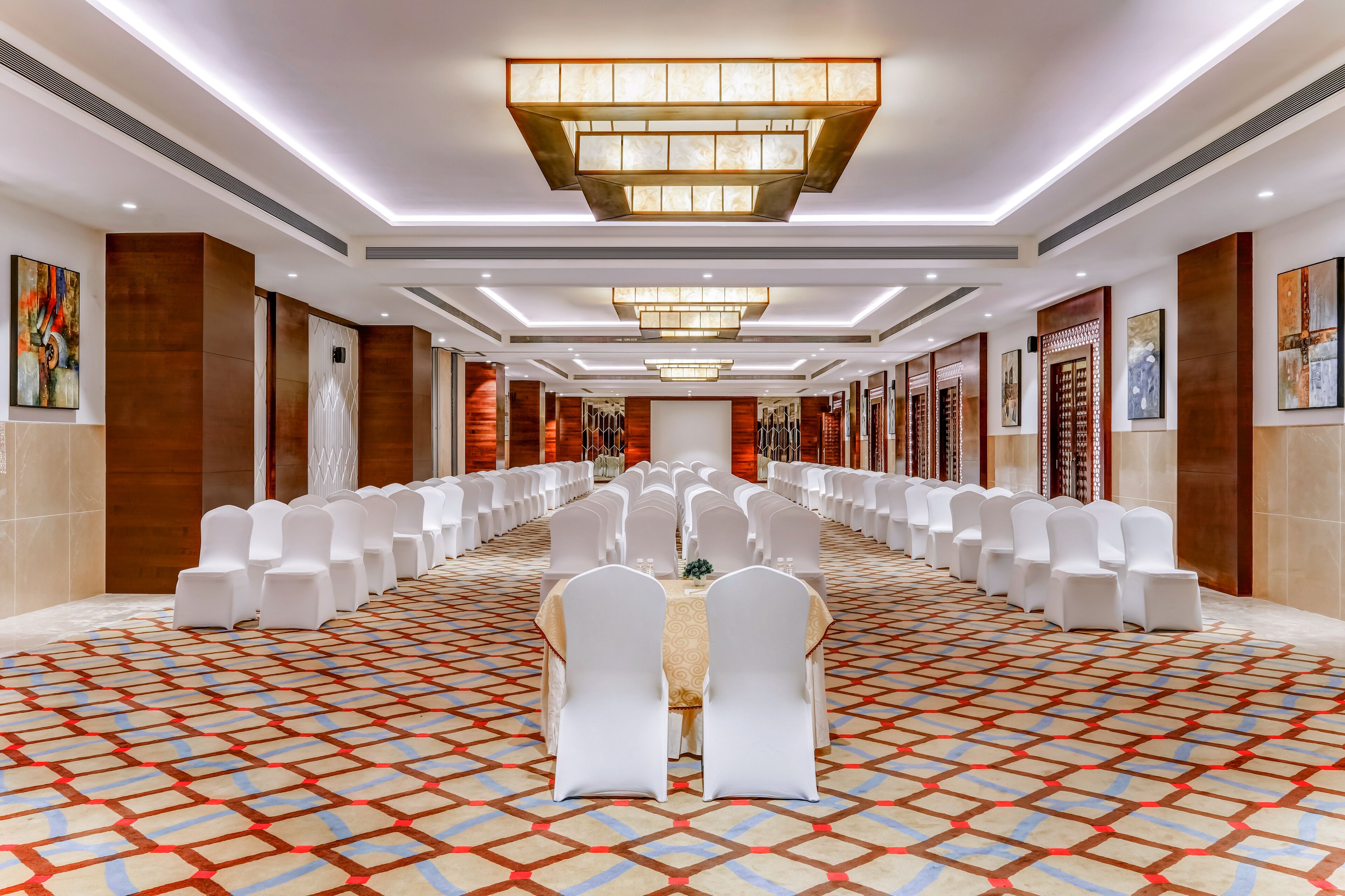 Um salão de banquetes extenso, com fileiras de cadeiras e mesas brancas, piso estampado e luminárias modernas.