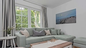 En moderne stue med en grå hjørnesofa, et træ sofabord og en lille rund fodsæde. Der er et billede af en bro på væggen og en potteplante på et sofabord.