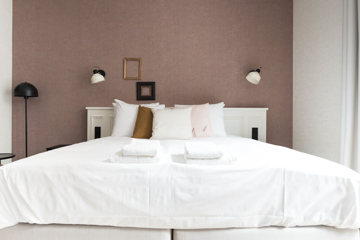 Una cama bien hecha con sábanas blancas y algunos almohadones, una lámpara de noche y un cuadro en la pared.