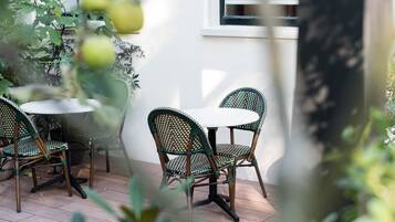 Un patio con sillas de mimbre y una mesa redonda, rodeado de plantas en macetas y una pared blanca.