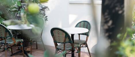 Un patio con sillas de mimbre y una mesa redonda, rodeado de plantas en macetas y una pared blanca.