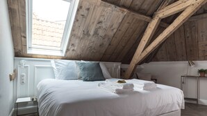Une chambre avec un plafond en pente, un lit recouvert de draps blancs, une fenêtre et une table de chevet avec une lampe.