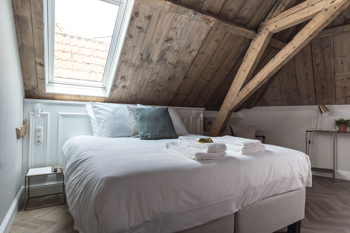 Un dormitorio con techo inclinado, una cama con ropa blanca, una ventana y una mesita de noche con lámpara.