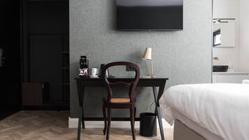 A hotel room with a bed, a desk with a chair, a television mounted on the wall, and a small trash bin.