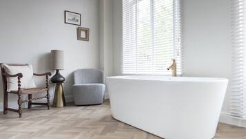 A modern bathroom with a freestanding bathtub, a wooden chair, a grey armchair, and a floor lamp.