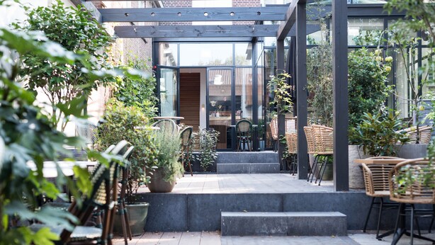 Un espace extérieur aménagé en patio, avec des meubles pour s’asseoir, des plantes en pot et une entrée vitrée menant à un bâtiment.