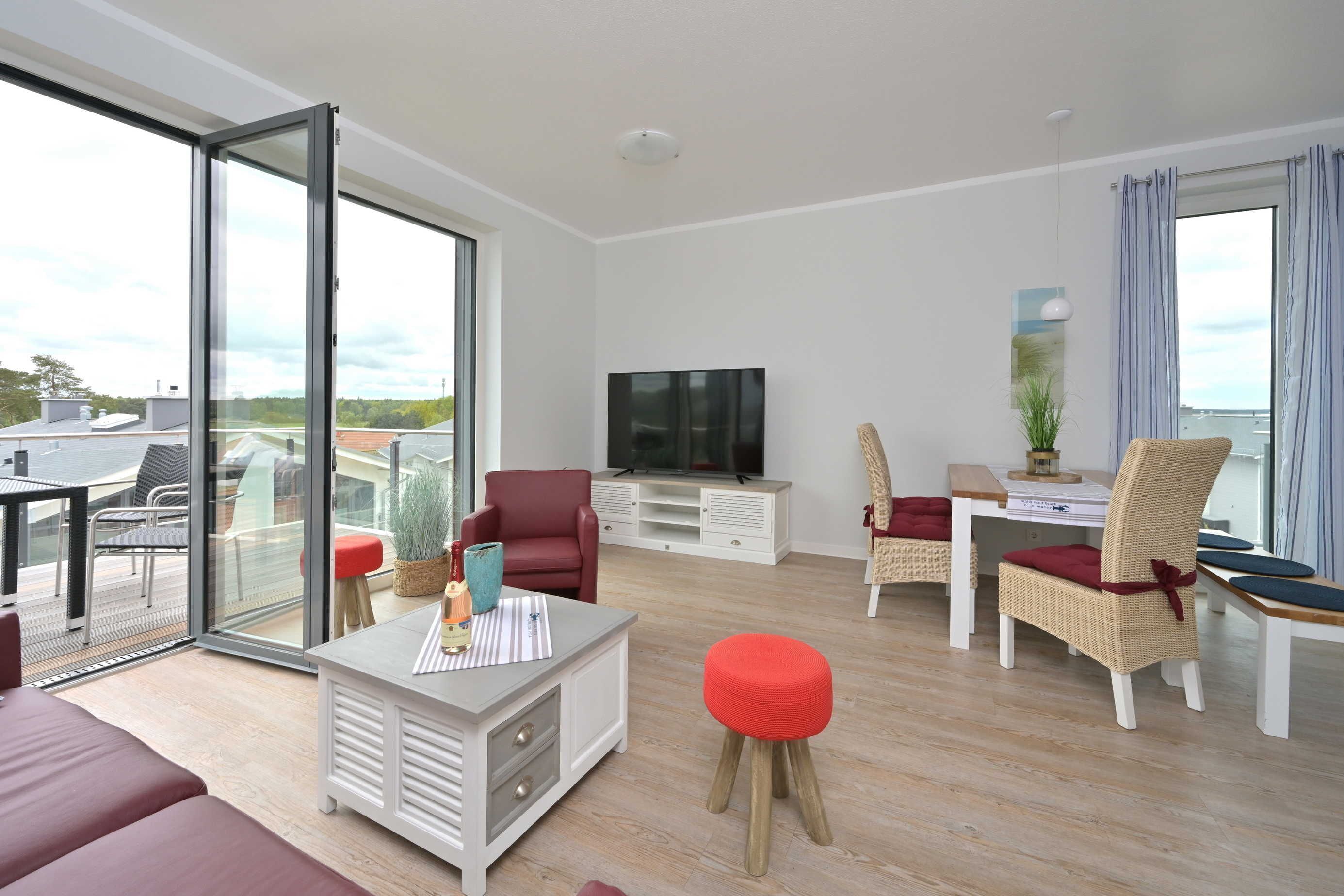 Una sala moderna con televisor de pantalla plana, comedor con sillas de mimbre y un amplio ventanal corredero que da a una terraza exterior.