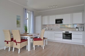 A modern kitchen with white cabinets, a microwave, and an oven. There is a dining table with wicker chairs and red cushions. A plant is on the table, and a painting hangs on the wall. - BoddenPanorama, Germany (Glowe)