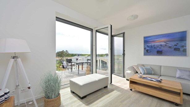 A modern living room with a large sliding glass door leading to an outdoor patio, a white sofa, a wooden coffee table, and a tripod lamp.