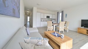 A modern living room with a sofa, coffee table, and a TV. There is a dining area with chairs and a table, and a kitchen with white cabinets and a microwave.