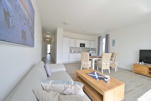 Un salon moderne avec un canapé, une table basse et un téléviseur. On y trouve également un coin repas avec des chaises et une table, ainsi qu’une cuisine dotée de meubles blancs et d’un four à micro-ondes.