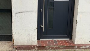 A black door with a metal handle and a glass panel, a small sign on the wall, and a brick base. - City Maritim Rooms (Bremerhaven)