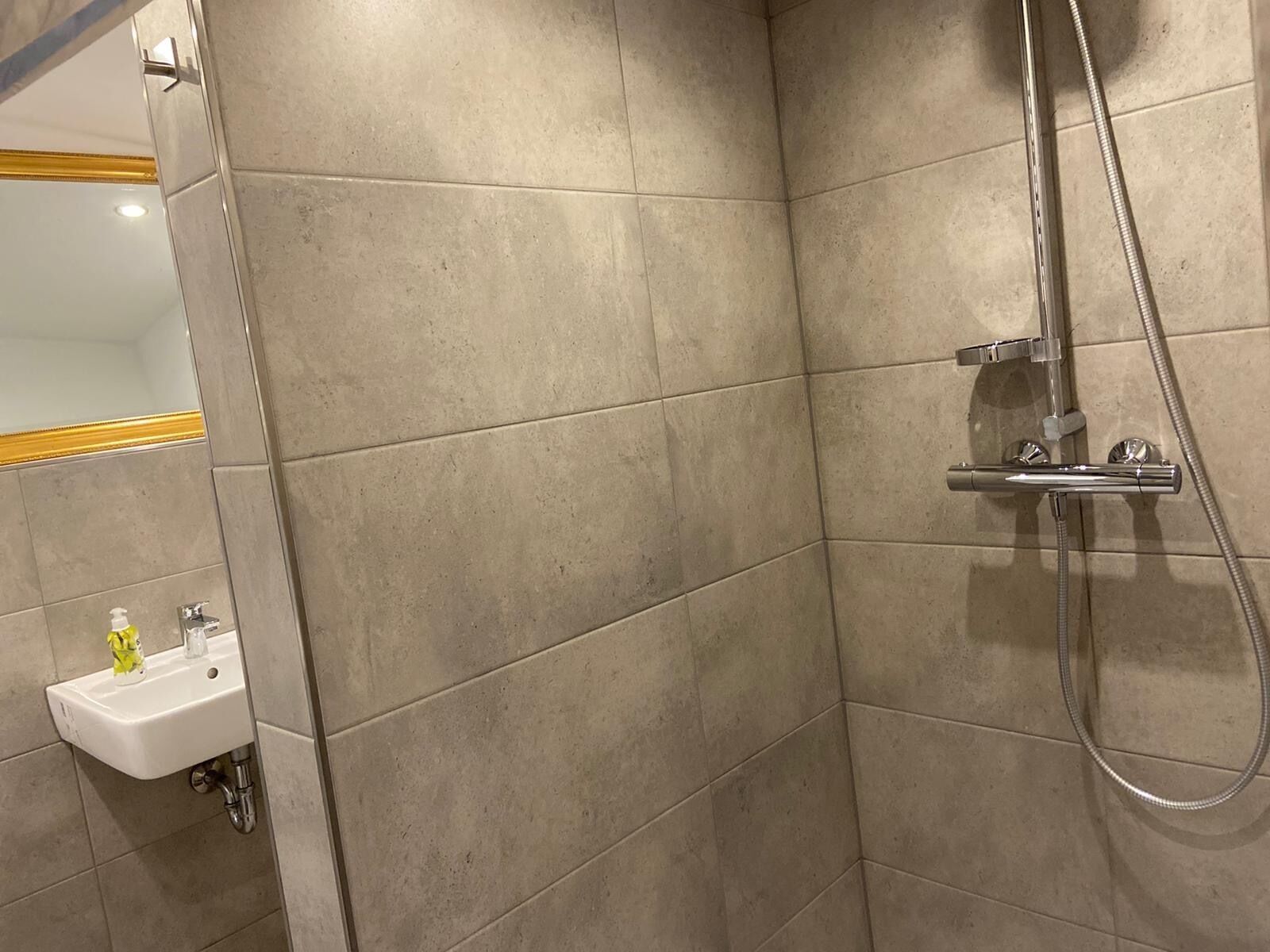 A bathroom with beige tiles, a shower with a handheld hose, and a sink with a bottle of hand soap.