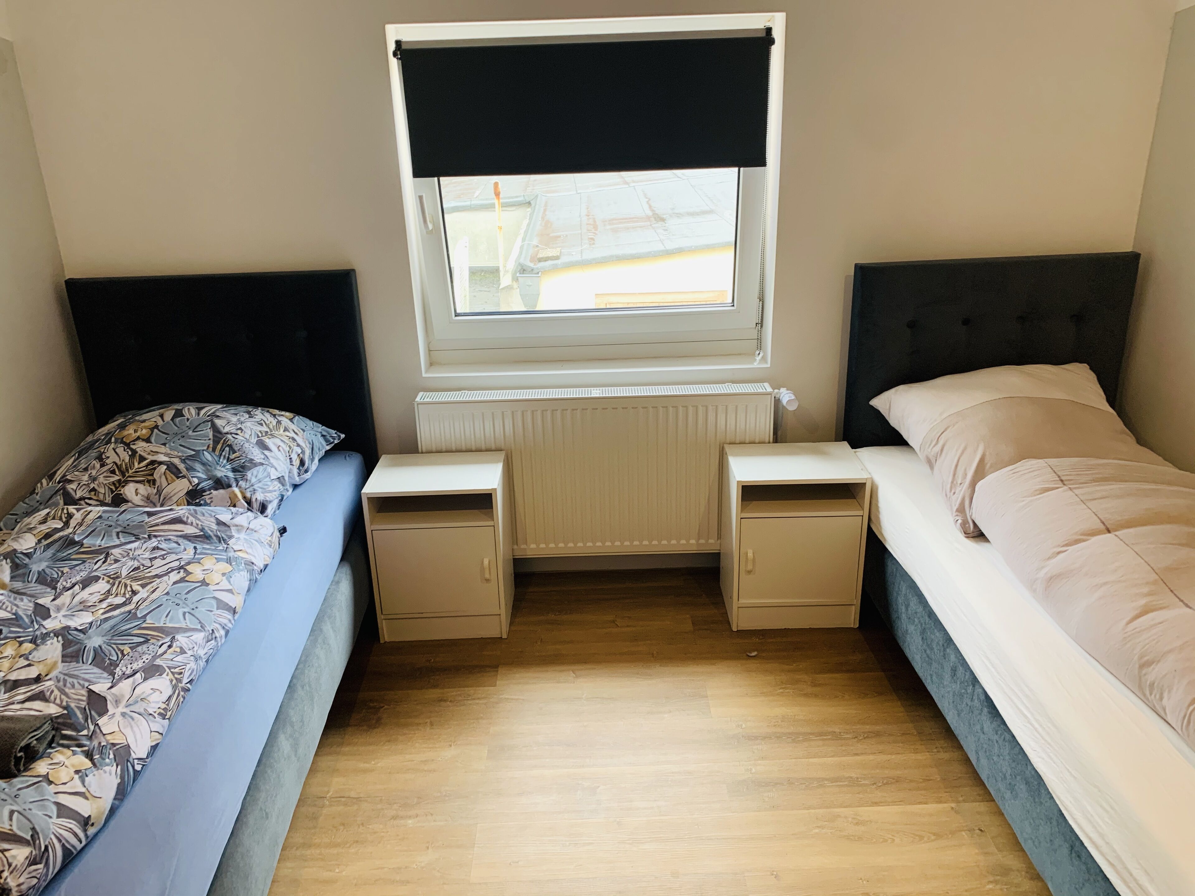 Een kleine kamer met twee bedden, een raam met een donkerblinde, een radiator en een houten vloer.