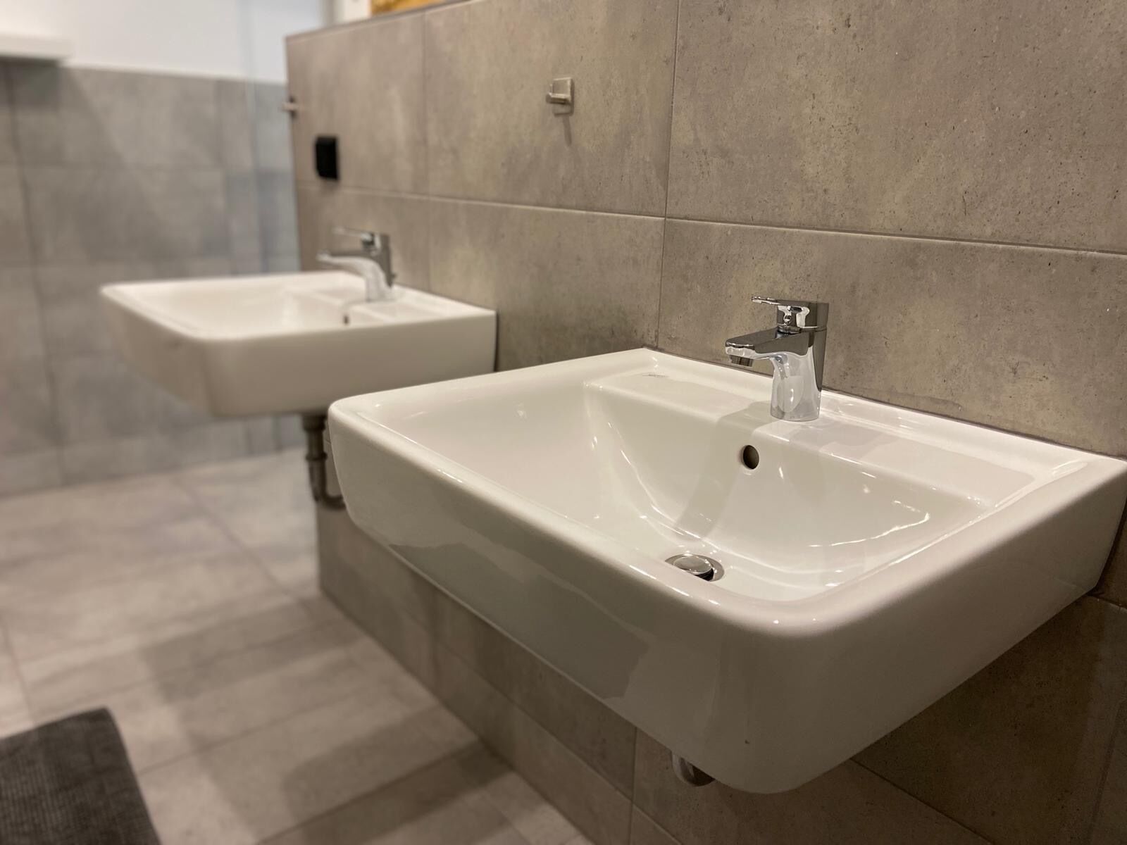Ein modernes Badezimmer mit zwei weißen Waschbecken an einer gefliesten Wand, einem sichtbaren Wasserhahn und einer kleinen Delle im Waschbecken.