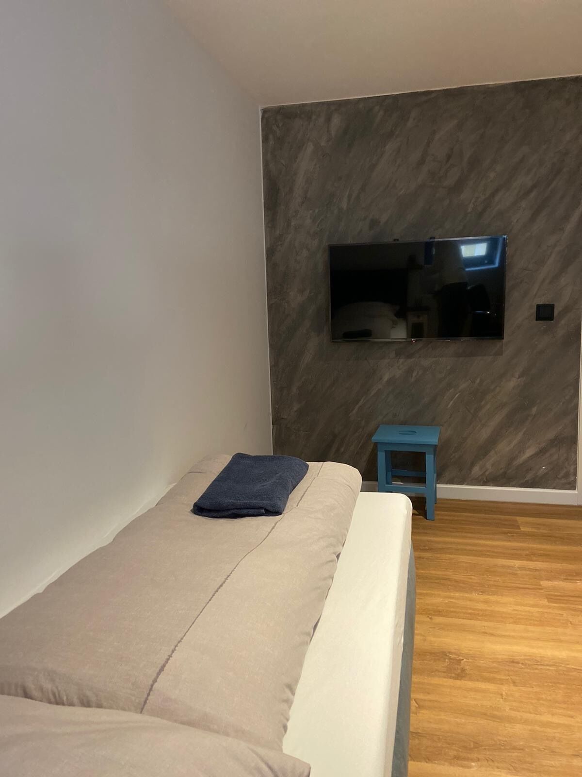 A modern bedroom with a bed, a small blue stool, a flat-screen TV mounted on the wall, and wooden flooring.