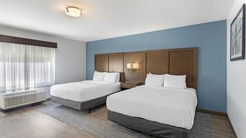 Quarto de hotel com duas camas, uma janela grande com cortinas leves, uma luminária na parede e um lustre no teto.